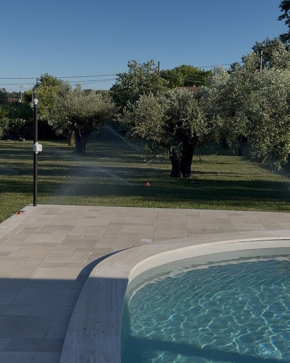 Irrigatori in funzione fra gli ulivi al tramonto, con il getto che attraversa il prato accanto al bordo della piscina
